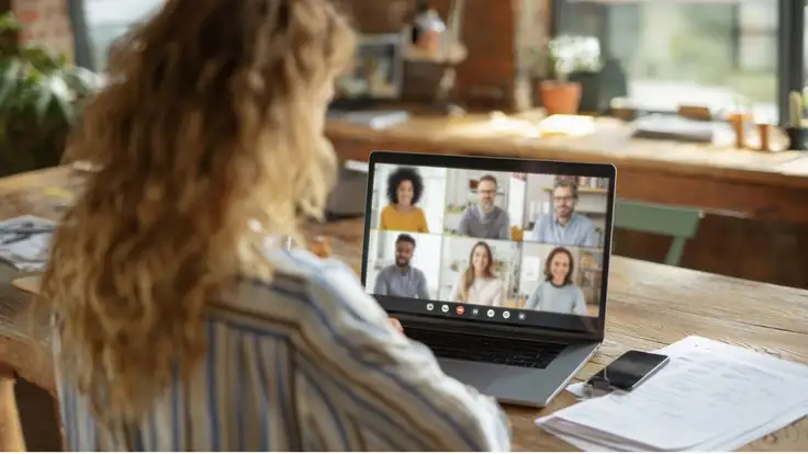 Vista desde atrás de una mujer en una oficina teniendo una videollamada grupal con seis colegas en su laptop sobre una mesa de madera, representando una sana cultura organizacional. 