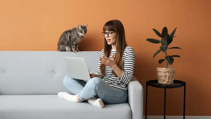 Chica estudiante sentada en un sofá con una laptop y un gato, lista para participar en clases virtuales con confianza. 
