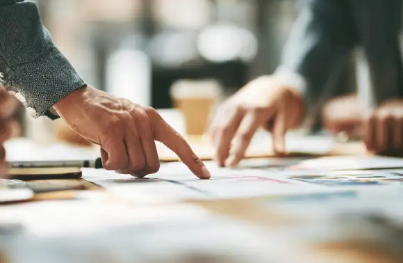 Personas revisando y señalando documentos impresos en una mesa durante una reunión de trabajo sobre impacto social y económico.