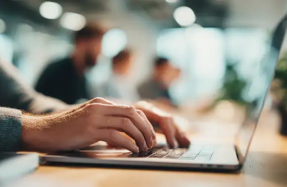 Manos de un pprofesional joven en un entorno colaborativo de oficina, destacando frente a un equipo, simbolizando la evolución de la publicidad hacia enfoques más humanos y digitales.