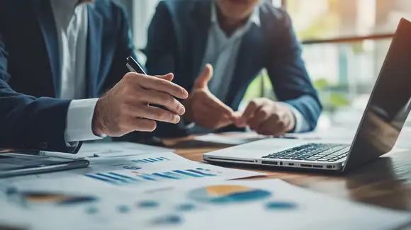 Dos profesionales en una oficina frente a una laptop conversan y analizan información y logística, reflejando colaboración y trabajo en equipo.