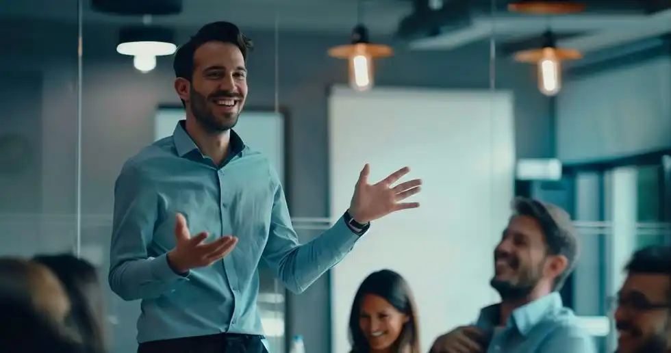 Foto de un instructor dentro de una oficina dando una charla de pie a sus compañeros de trabajo sobre bienestar personal en beneficio de la productividad laboral.