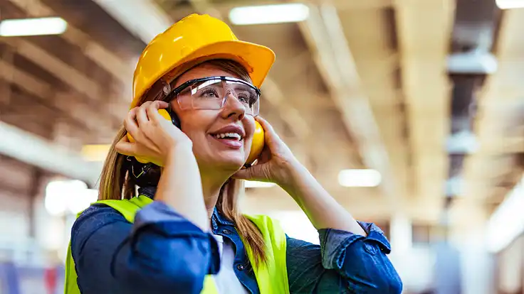 Mujer con chaleco de seguridad y protectores auditivos, sentada en un almacén industrial, representando prácticas de salud ocupacional y promoción de la salud en el trabajo.